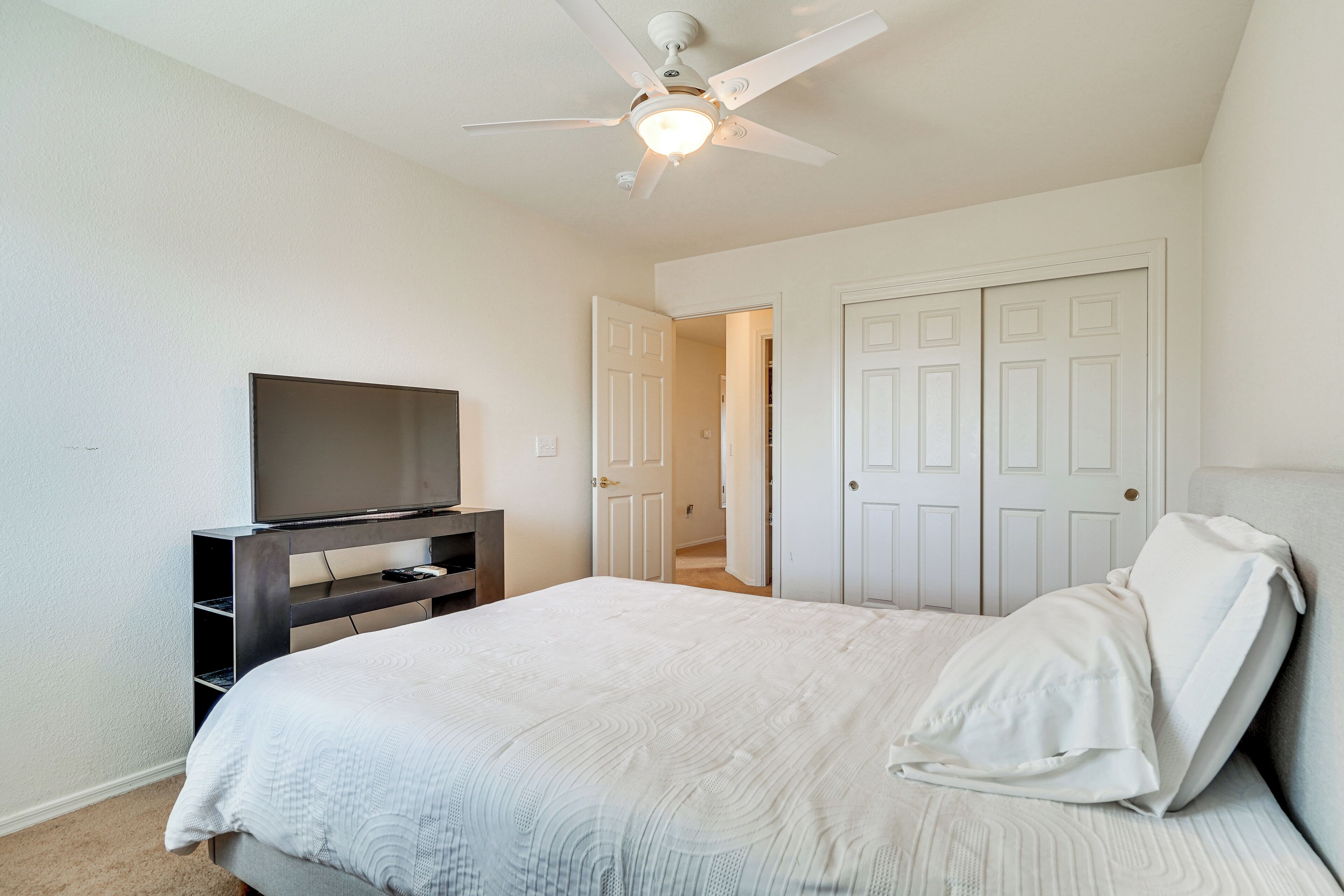 A bedroom with a bed, a TV on a stand, a ceiling fan, and a door leading to another room.
