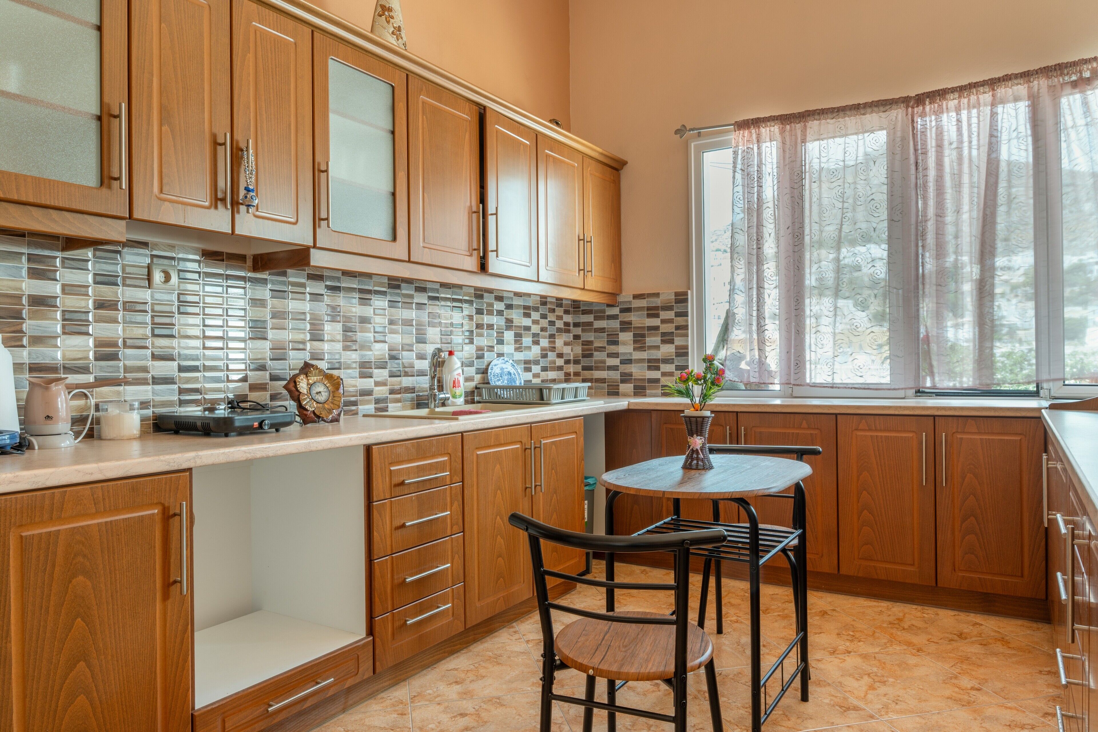 A kitchen with wooden cabinets, a tiled backsplash, a coffee maker, a kettle, and a window with curtains.