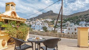 A terrace with two wicker chairs and a table, overlooking a town with white buildings and a hill in the background.