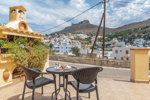 A terrace with two wicker chairs and a table, overlooking a town with white buildings and a hill in the background.