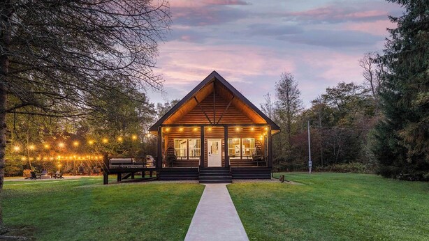 A wooden cabin with a porch, a pathway leading to the entrance, and string lights. - Aviation Cabin | Hot Tub • Fire Pit • River Access (Roulette)