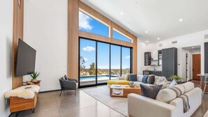 A spacious living room with a large window overlooking a pool, a flat-screen TV mounted on the wall, and a modern kitchen area. - The Mesa Horizon House By Homestead Modern (Yucca Valley)