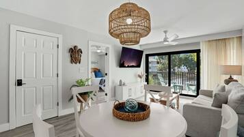 A modern living room with a round white table, a wicker chandelier, a grey sofa, and a large window with a view of the outdoors.