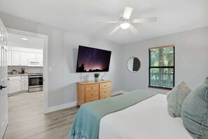 A bedroom with a bed, a ceiling fan, a TV on the wall, a window with blinds, and a view of the kitchen.