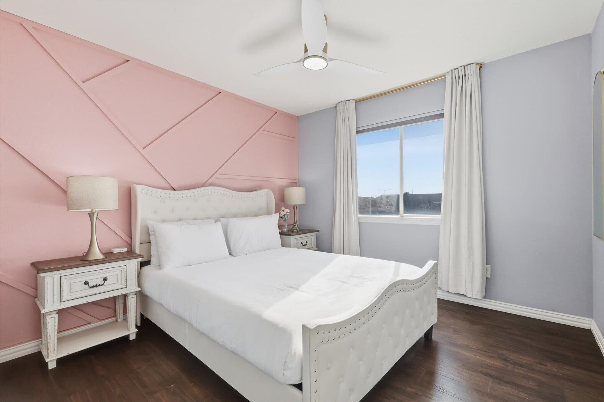 Une chambre à coucher avec un lit, des tables de chevet, un ventilateur de plafond, une fenêtre avec des rideaux et une vue sur la ville.