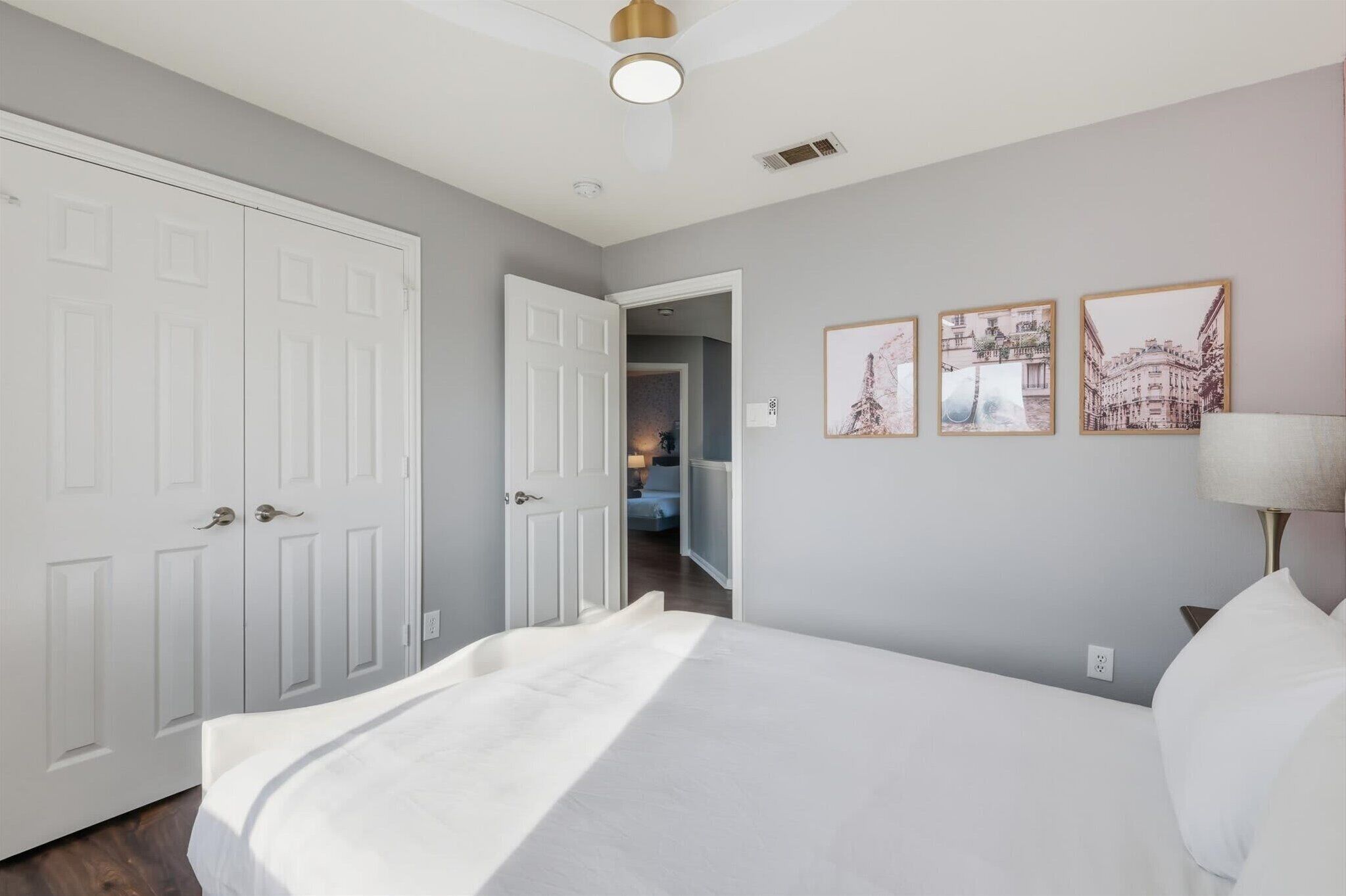 A bedroom with a bed, white walls, and a door leading to another room. There are three framed pictures on the wall.