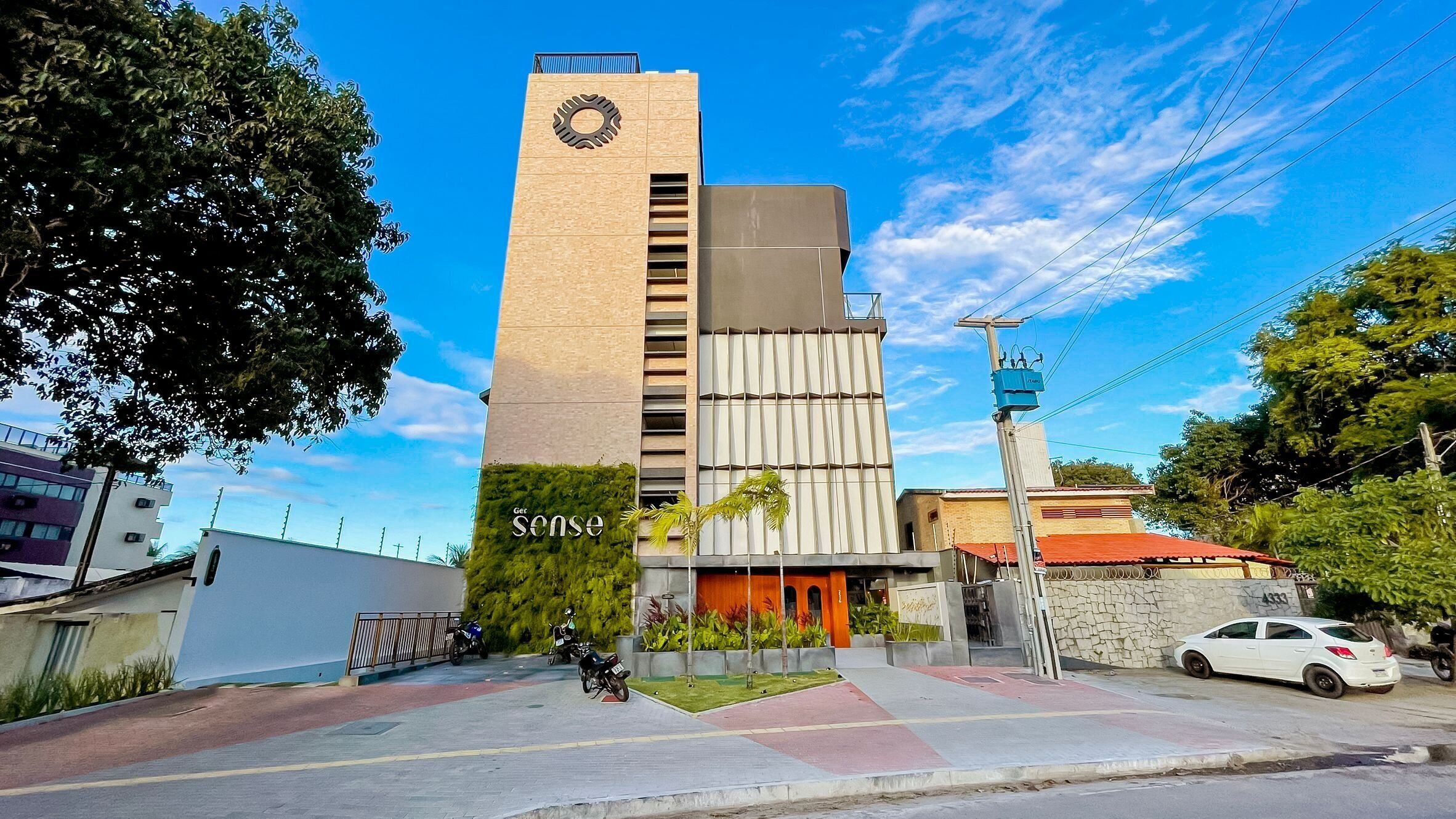 Un edificio moderno con una destacada torre de reloj, una entrada portonada y un estacionamiento con motocicletas y automóviles.