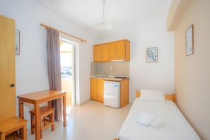 Quarto compacto com cama, uma mesinha, área de cozinha com armários e geladeira, e uma porta que dá para uma área externa.