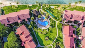 An aerial view of a resort with multiple red-roofed buildings, a central swimming pool, and surrounding palm trees. - Papakea Ocean View Resort Condo | Sleeps 7, Pools, Hot Tubs, Tennis, Lanai, AC (Lahaina)