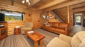 Interior d'una cabana de fusta amb un paviment de fusta, un sofà de cuir marró, un butac armchair beix, una taula de centre de fusta, una televisió i una escala que condueix a una altra habitació.