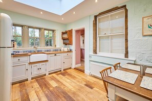Una cocina con piso de madera, armarios blancos, un ventanal con persianas y una zona de comedor con una mesa y sillas.