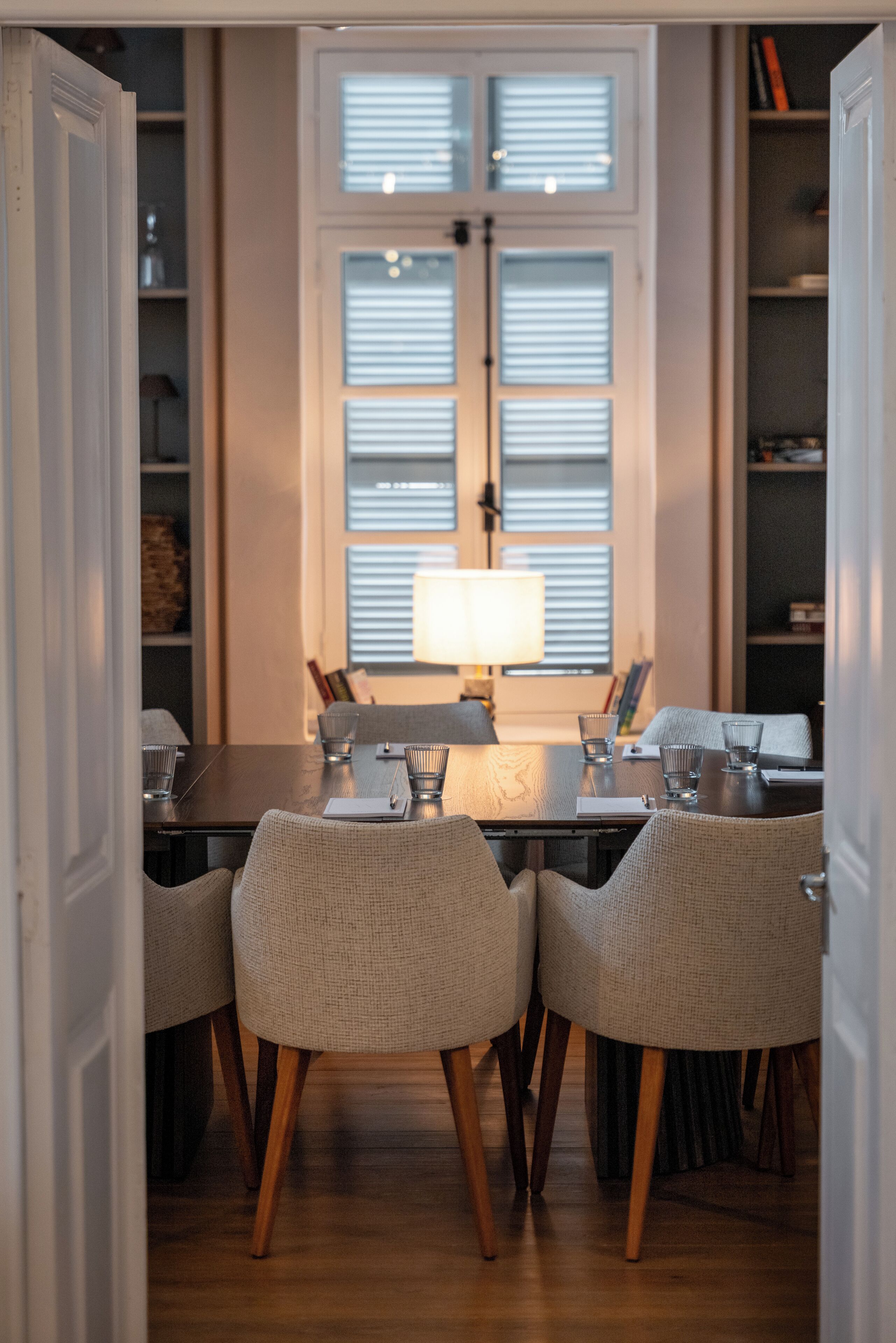 A dining area with a table set for four, flanked by wicker chairs, a lamp, and a window with blinds.