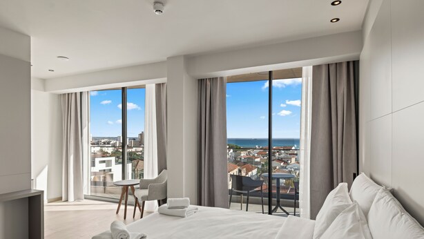 A modern hotel room with a large bed, a small table, and a chair. The room has large windows with a view of the city and the sea. - Warner Boutique Hotel (Larnaca)