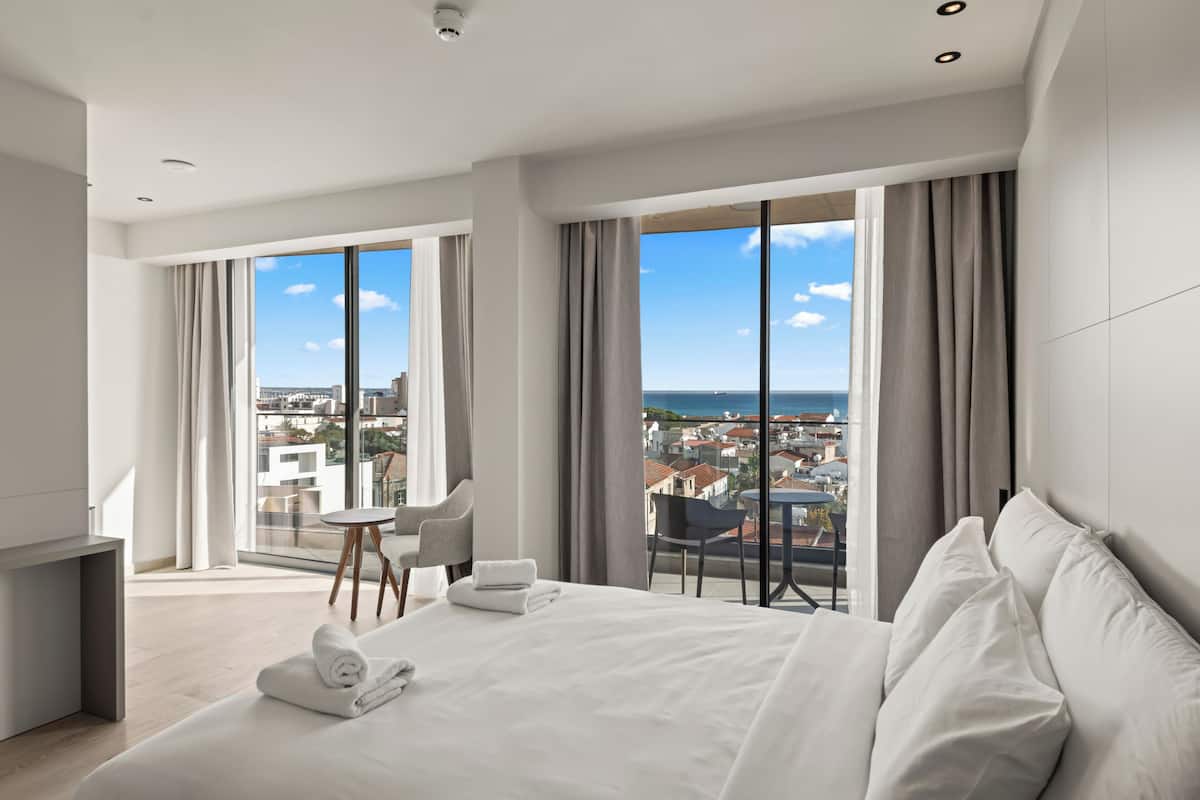 Habitación de hotel moderna con una cama grande, una mesita pequeña y una silla. La habitación tiene ventanas amplias con vistas a la ciudad y al mar.