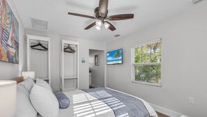 A bedroom with a bed, a ceiling fan, a TV, a window with a view of palm trees, and a door leading to a bathroom. - 🌞400 ft to the Beach👙Onsite Heated Pool🐬Bungalows #2🌴Grills🐶Dogs Welcome! (Treasure Island)