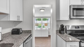 Une cuisine moderne avec des meubles blancs, des plans de travail en granit, un four encastré et une fenêtre équipée de stores.