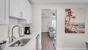 Cocina moderna con armarios blancos, un lavamanos de acero inoxidable y una cafetera. Cuenta con un comedor con mesa y sillas, y un cuadro de palmeras en la pared.