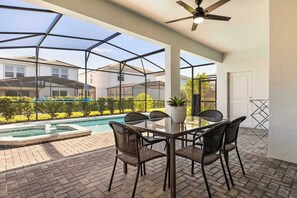 Une terrasse extérieure avec une table et des chaises en verre, un ventilateur de plafond, et une vue sur une piscine et des maisons en arrière-plan.