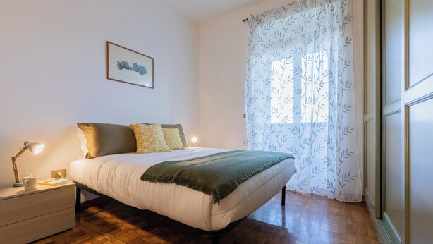A bedroom with a bed, bedside table, lamp, and a window with patterned curtains. - Casa Cecchi by Wonderful Italy (Turin)