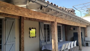 A covered outdoor seating area with a table and chairs, a wooden post, and a door with a window. - Les Berbéris – Gîte in Southern Ardèche (Chandolas)