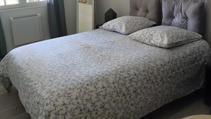 A bedroom with a bed, a mirror, a radiator, a window with curtains, and a wall-mounted artwork. - Les Berbéris – Gîte in Southern Ardèche (Chandolas)