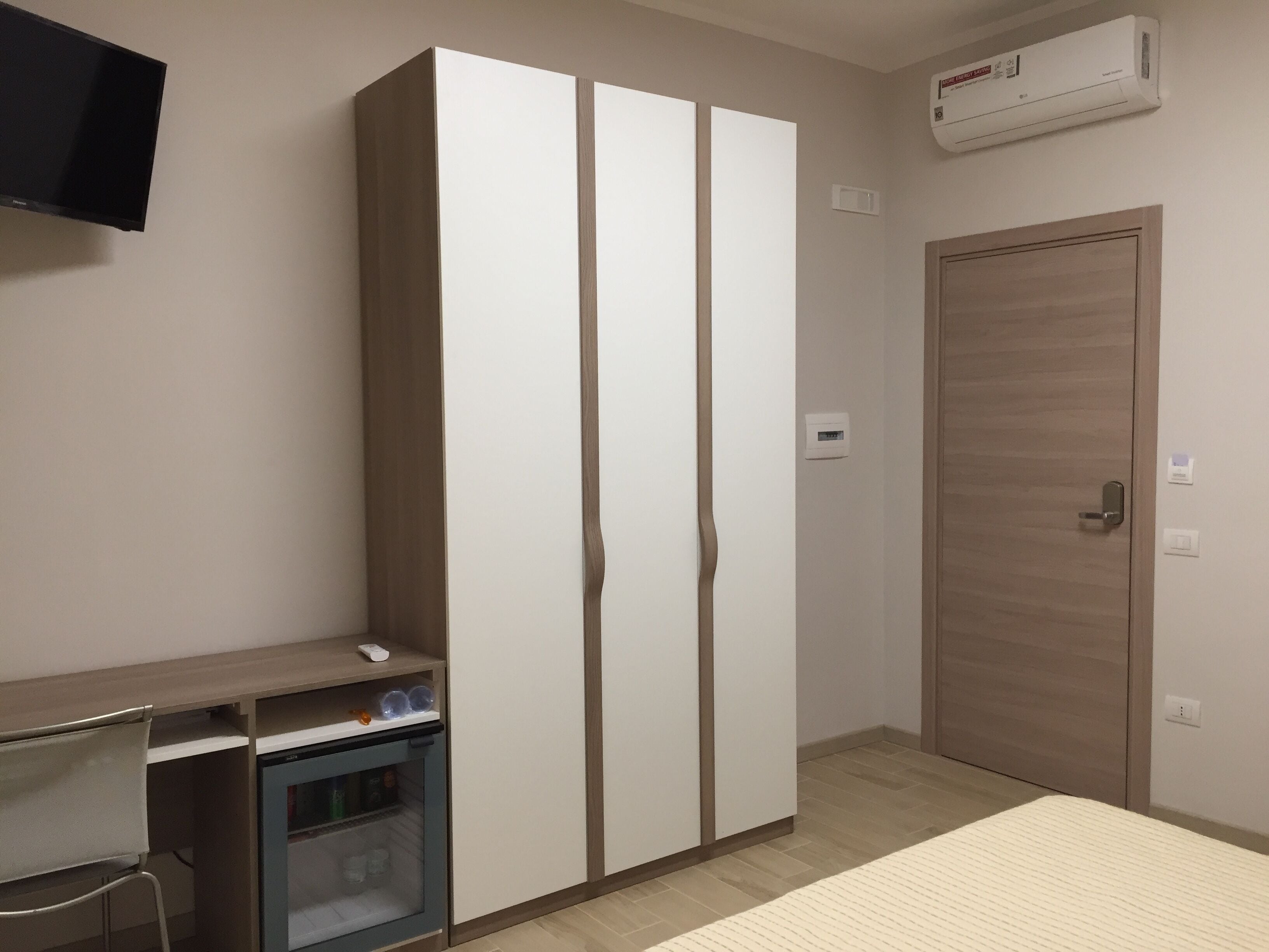 A bedroom with a wardrobe, a desk with a mini-fridge, and an air conditioning unit.