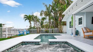 Een zwembad met een jacuzzi, omringd door een witte hek, palmbomen en een huis met een overdekt terras.