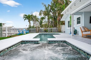 A swimming pool with a hot tub, surrounded by a white fence, palm trees, and a house with a covered patio.