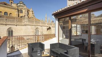 Un balcon agrémenté de meubles en osier, une porte vitrée donnant sur un espace de restauration intérieur, et un bâtiment historique à l’architecture riche en détails en arrière-plan.
