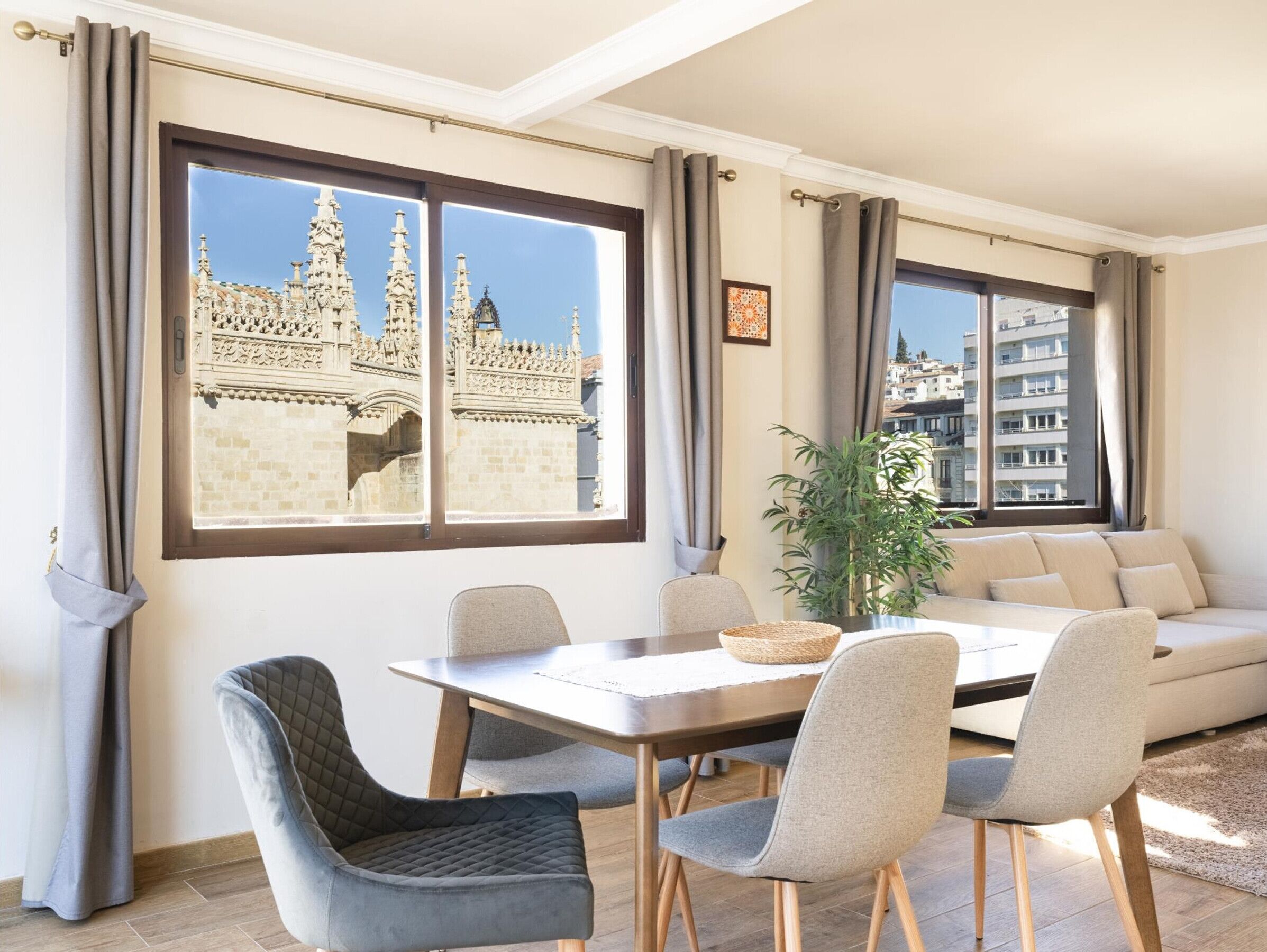 Una sala de estar moderna con comedor, ventanas amplias con vista a un edificio histórico y una planta en maceta.