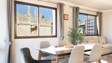 A modern living room with a dining area, large windows with a view of a historic building, and a potted plant.
