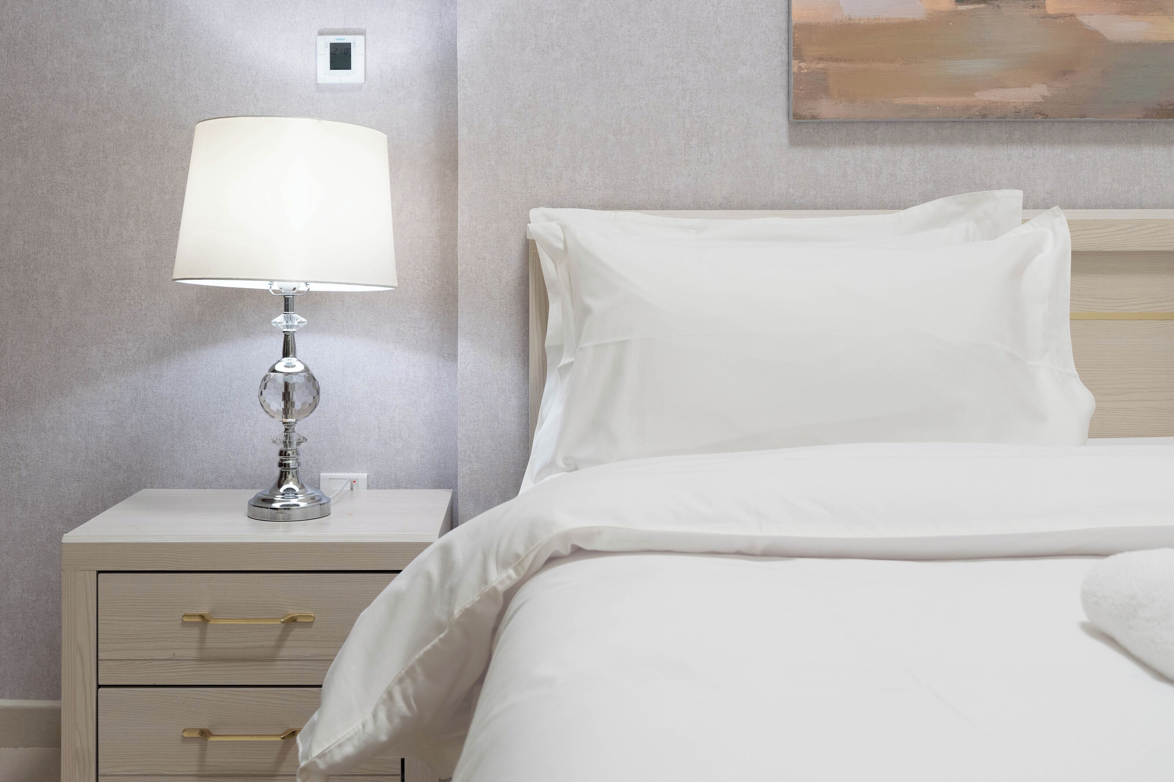 Een net opgemaakt bed met witte lakens, een bedlampje en een nachtkastje met lades.