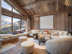 A cozy living room with a large window, a sofa, ottomans, and a coffee table. - Modern chalet in Courchevel with sauna and jacuzzi (Courchevel 1550)
