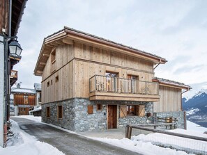 A two-story wooden chalet with a stone foundation, a balcony with metal railings, and a view of snow-covered mountains in the background. - Modern chalet in Courchevel with sauna and jacuzzi (Courchevel 1550)