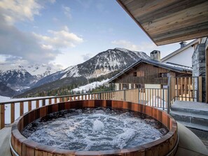 A hot tub with a wooden exterior, set against a backdrop of snow-covered mountains and a wooden building with a balcony. - Modern chalet in Courchevel with sauna and jacuzzi (Courchevel 1550)
