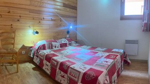 Ein gemütliches Schlafzimmer mit holzverkleideter Schräge, einem Bett mit einer rot-weißen Steppdecke, einem Holzstuhl, einem Wandheizkörper und einem Fenster mit Vorhängen.
