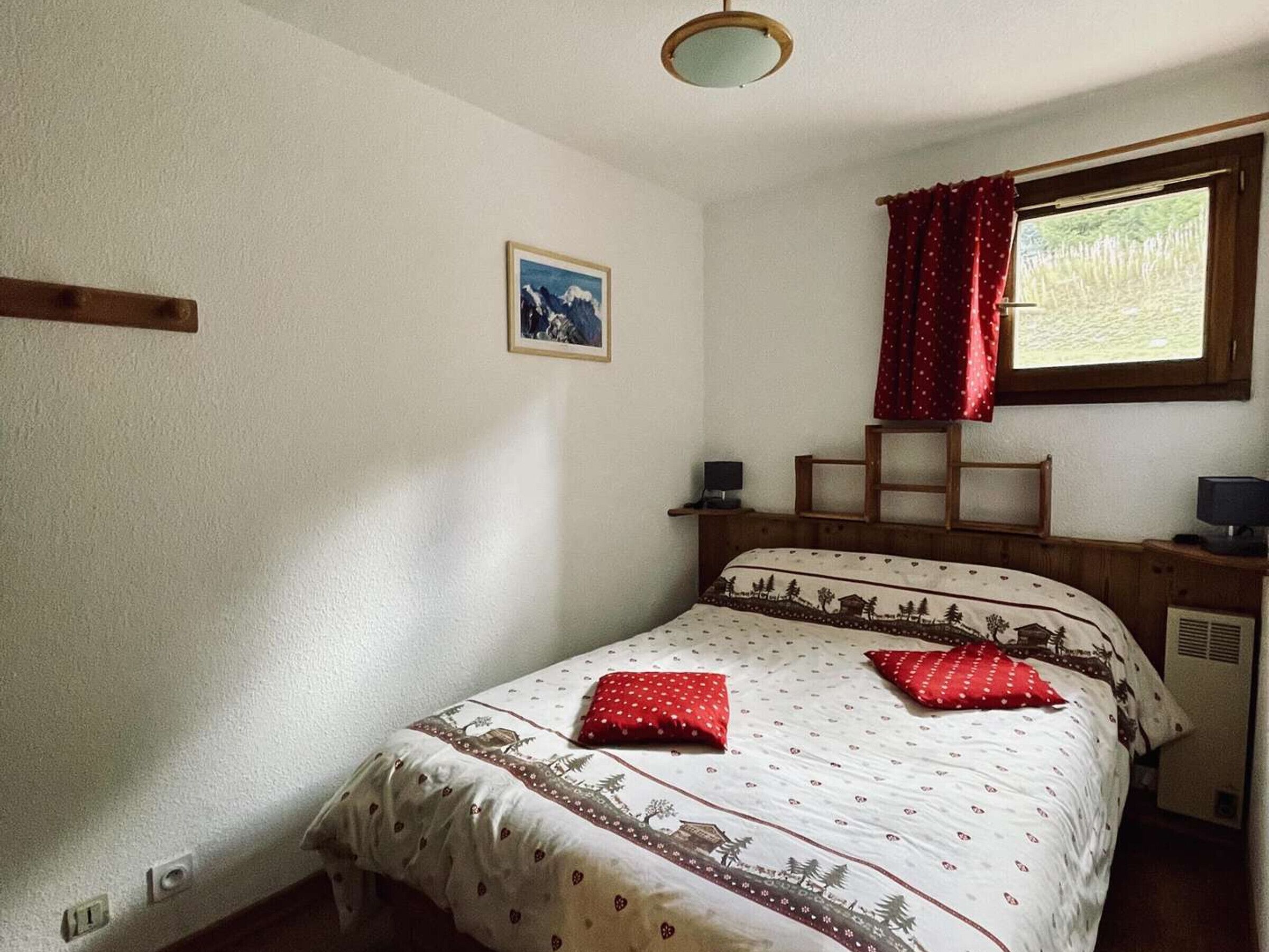 A bedroom with a bed, a small table, a chair, a window with curtains, a wall-mounted lamp, and a picture on the wall.