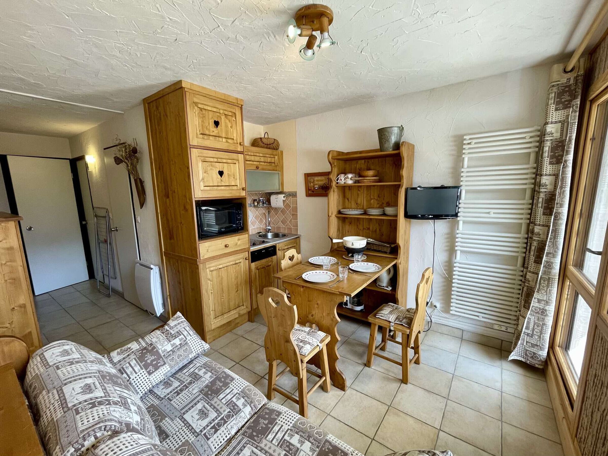 A compact kitchen with wooden cabinets, a dining table, and chairs. There is a built-in microwave and a small refrigerator.