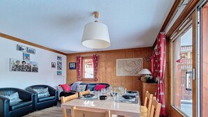 Un salón acogedor con una mesa de comedor de madera preparada para cuatro personas, un sofá azul y un amplio ventanal con cortinas.
