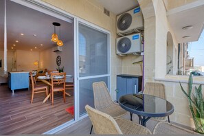 A dining area with wicker chairs and a glass table, a sliding glass door leading to a balcony, and air conditioning units mounted on the wall. - Qala Tranquil Escape - Stylish With Terrace AC (Qala)