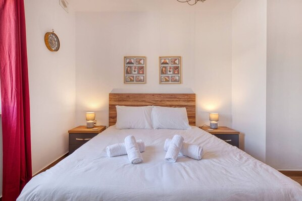 A neatly made bed with white linens and rolled towels, wooden headboard, two bedside tables with lamps, and framed pictures on the wall. - Qala Tranquil Escape - Stylish With Terrace AC (Qala)
