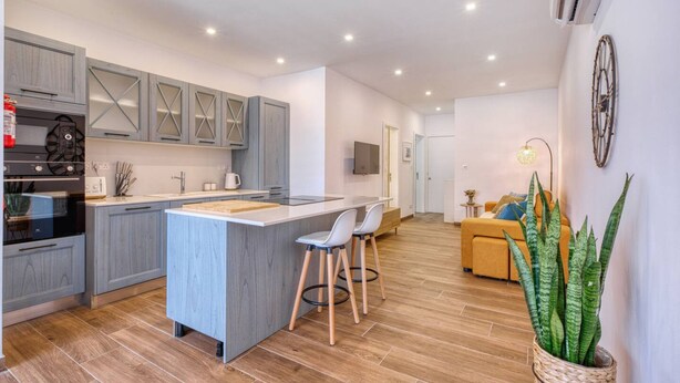 A modern kitchen with grey cabinetry, a central island with stools, and a dining area with a sofa and a plant. - Qala Tranquil Escape - Stylish With Terrace AC (Qala)