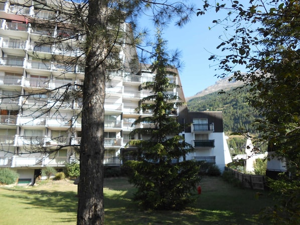A multi-story residential building with balconies, surrounded by trees and a mountain in the background.