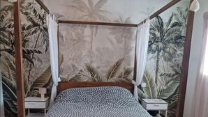 Un lit à baldaquin à quatre poteaux, agrémenté d’une couvre-lit à motifs et de deux oreillers, le tout placé devant un décor mural représentant un paysage tropical.