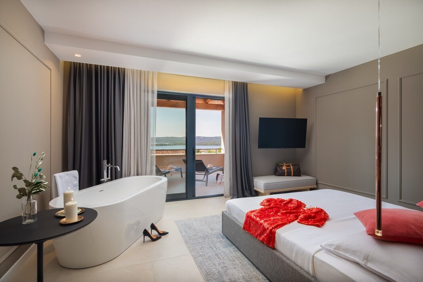 Ein modernes Hotelzimmer mit einer großen Badewanne, einem Bett mit roter Tagesdecke, einem Flachbildfernseher und einem Balkon mit Blick.