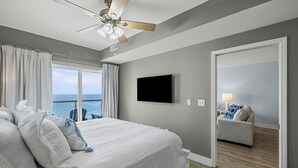 A hotel room with a bed, a ceiling fan, a TV, a view of the ocean, and a door leading to a balcony. - Gulf Front Upscale 2BR Condo | Sleeps 6 | 2 Pools | Hot Tub (Pensacola Beach)