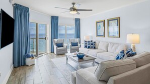 A modern living room with a large window, a ceiling fan, and a view of the ocean. - Gulf Front Upscale 2BR Condo | Sleeps 6 | 2 Pools | Hot Tub (Pensacola Beach)