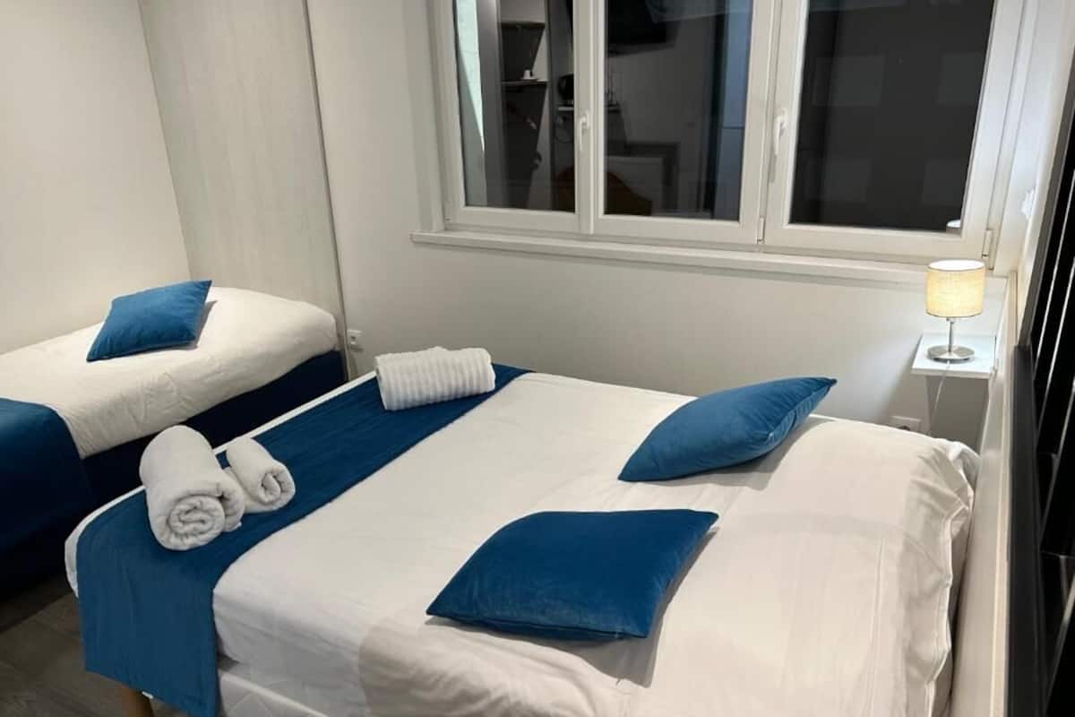 A hotel room with two beds, each with blue pillows and white linens. There is a window with blinds, a bedside table with a lamp, and a towel rack with towels.