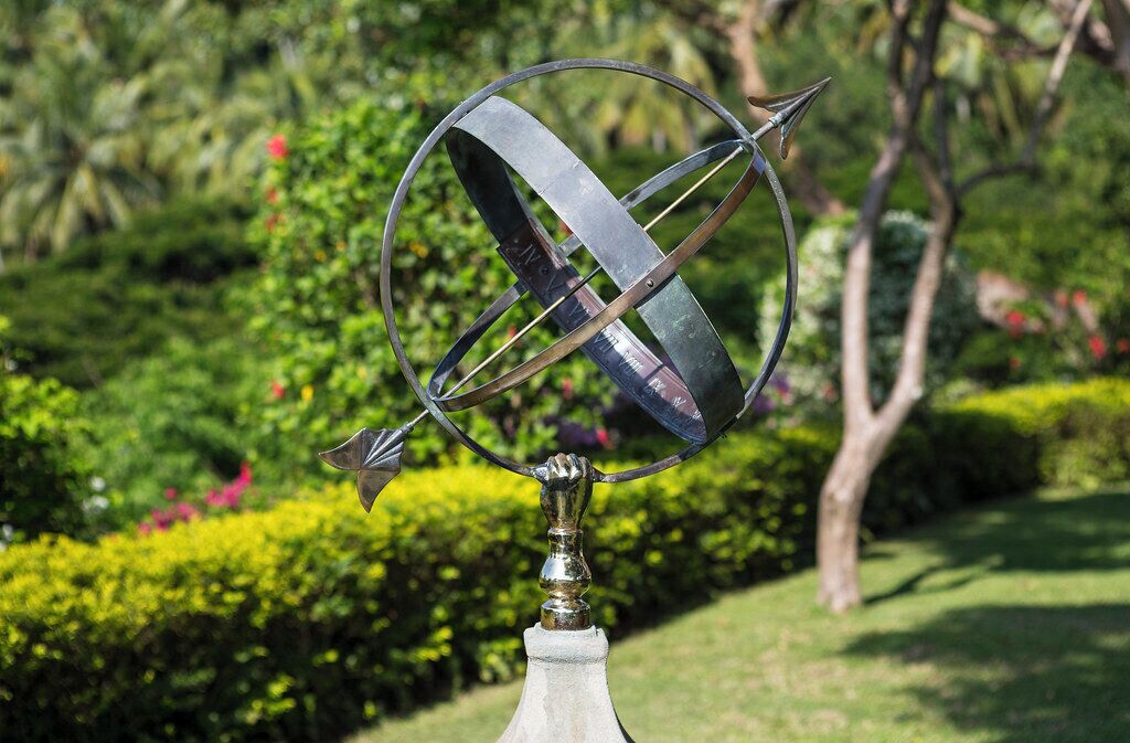 A metal sculpture with a circular base and intersecting elements, set against a lush garden backdrop.
