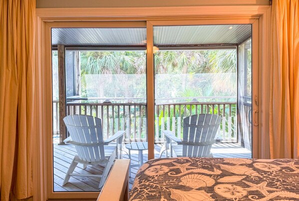 A room with a bed, a sliding glass door leading to an outdoor deck with two Adirondack chairs and a small table.